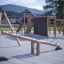 Lekestativ Seesaw Nature på en lekeplass med lekehus i bakgrunnen.