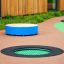Rund trampolin (100 cm) montert i lekeplassens gulv med fargerik gummibelegg rundt.