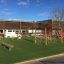 Lekeplass med Balansebom og klatrestativer i tre på et grønt, kunstgressdekke foran en skolebygning.